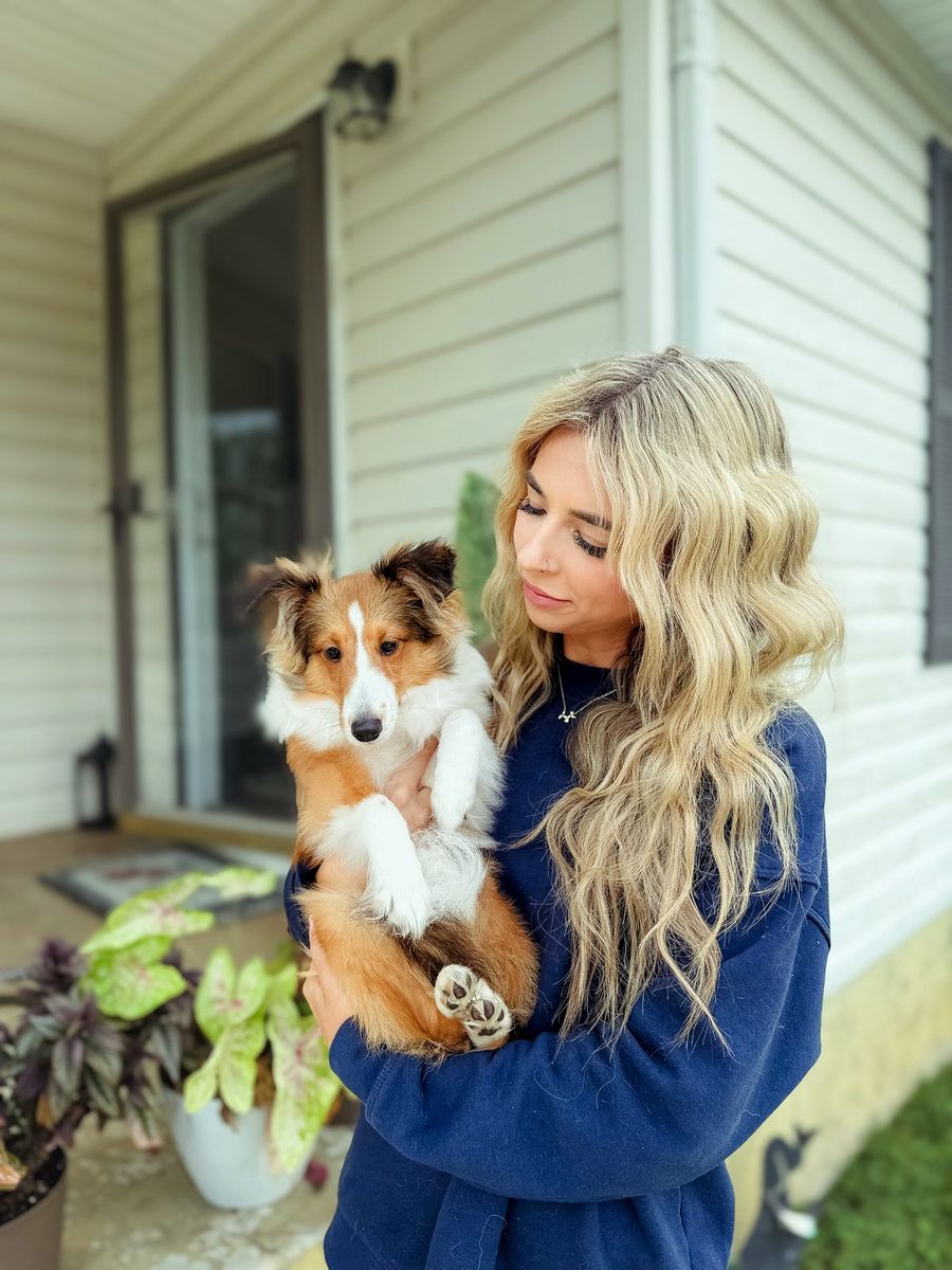 A trainer cradles a sheltie puppy on a front porch beside leafy plants
