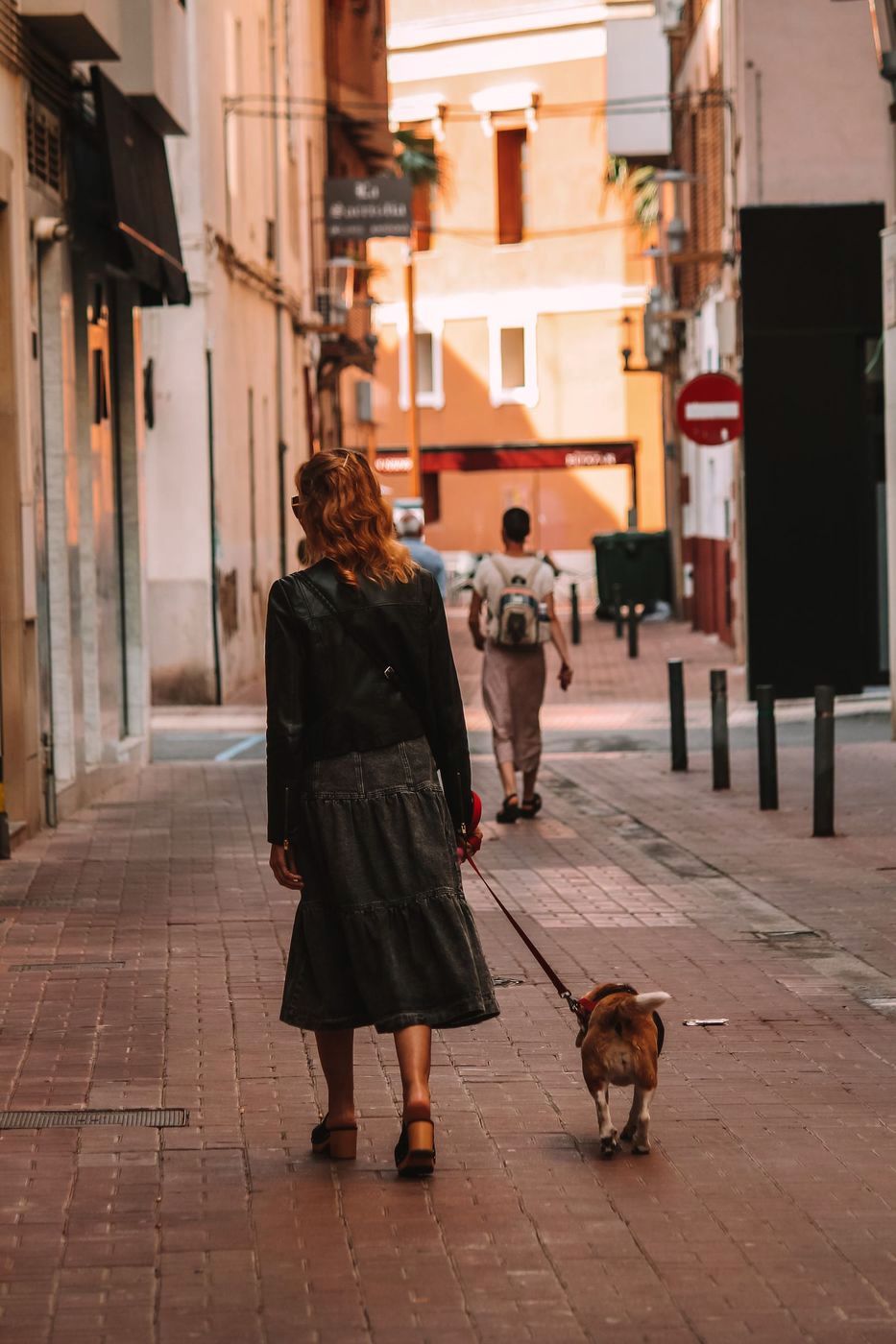 A woman walking a small dog down a sunlit old-town street